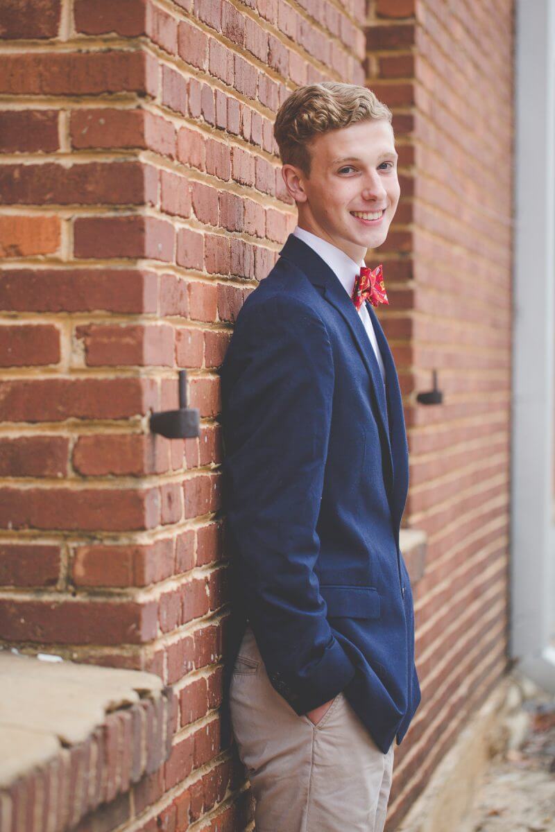 senior young man in sport coat posing with brick alley wall