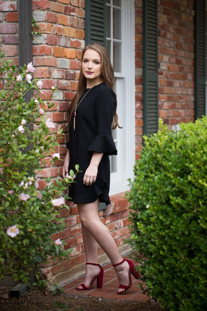 brunette high school senior in black dress and red heels posing in front of brick wall