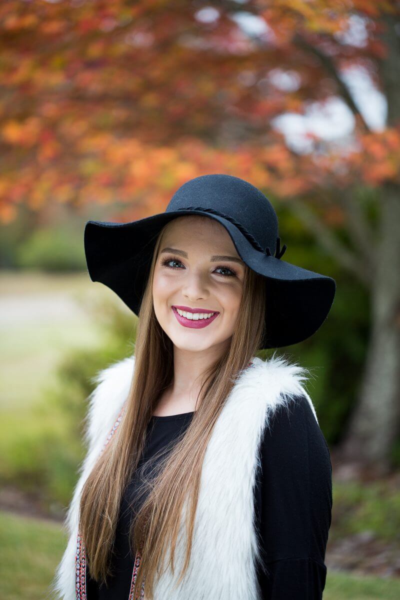 high school senior girl posing in black dress and hat under fall leaves