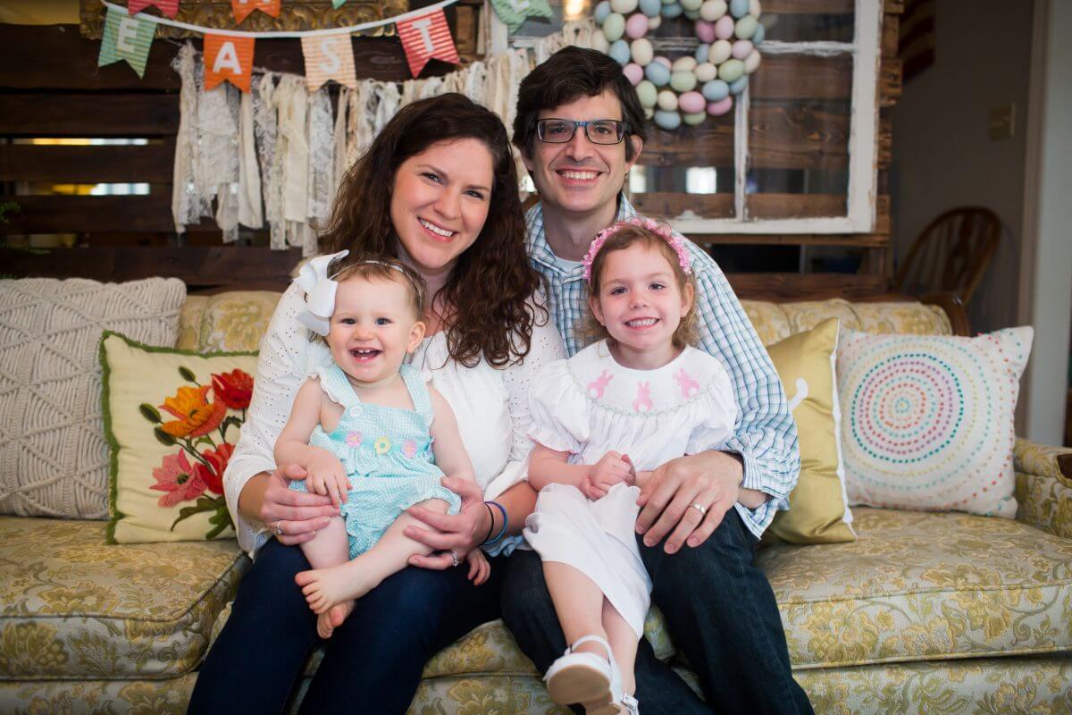 Family with young daughter and infant daughter pose for photo in Easter clothes on vintage couch