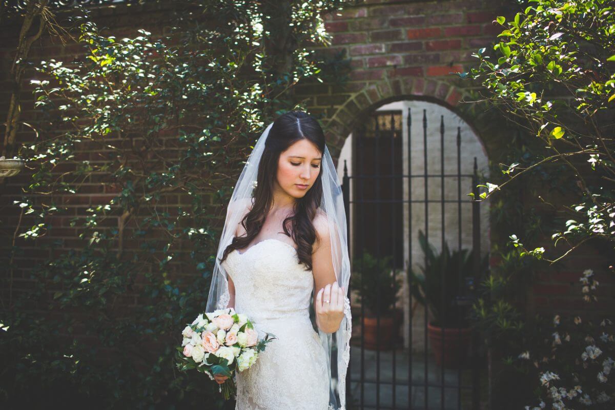 Demure bride inspecting veil in front of brick wall and garden gate