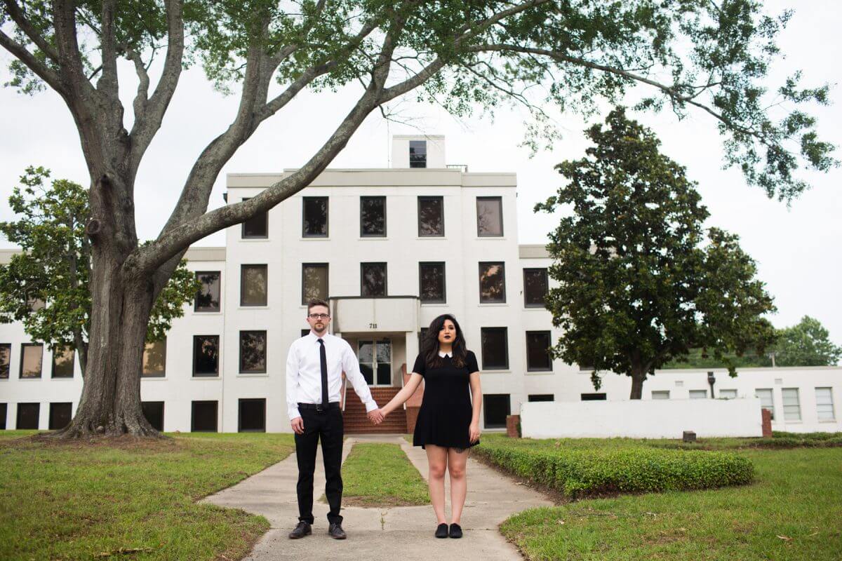 man and woman in black and white clothing holding hands in front of white abandoned building and large tree