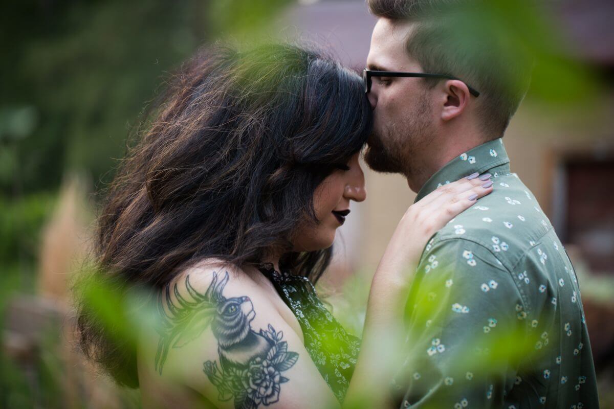 man in glasses kissing brunette woman with jackalope tattoo on forehead