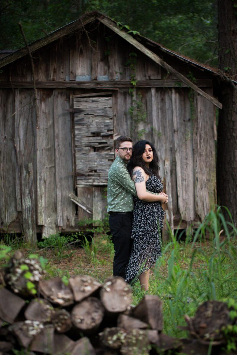 couple posing for engagement session in front of dilapidated wooden house with tin roof