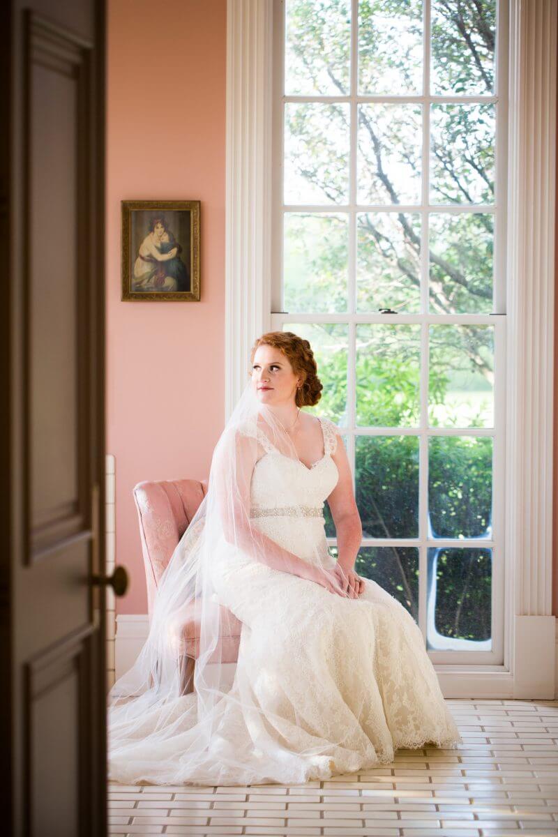 bride in veil sitting on pink chair in front of window