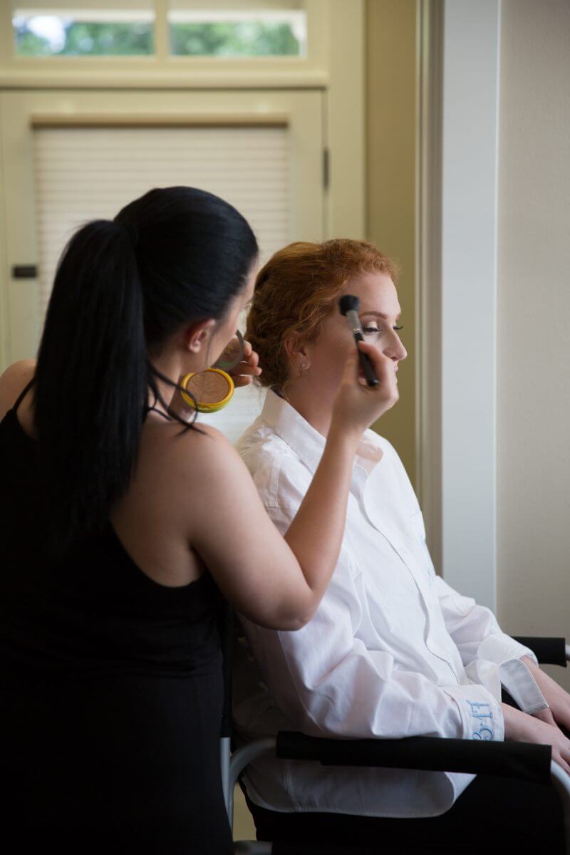 aesthetician applying makeup on redheaded bride in white robe