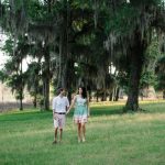 engaged couple holding hands and walk across green lawn under shady trees with Spanish moss