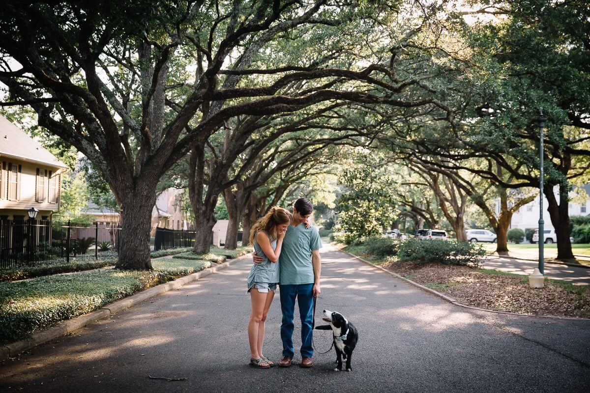 loving couple standing in an oak lined street and looking down at their dog who is looking up at them