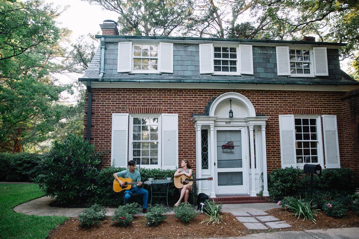 man and woman sitting with their guitars on the concrete walkway in front of their two story red brick home