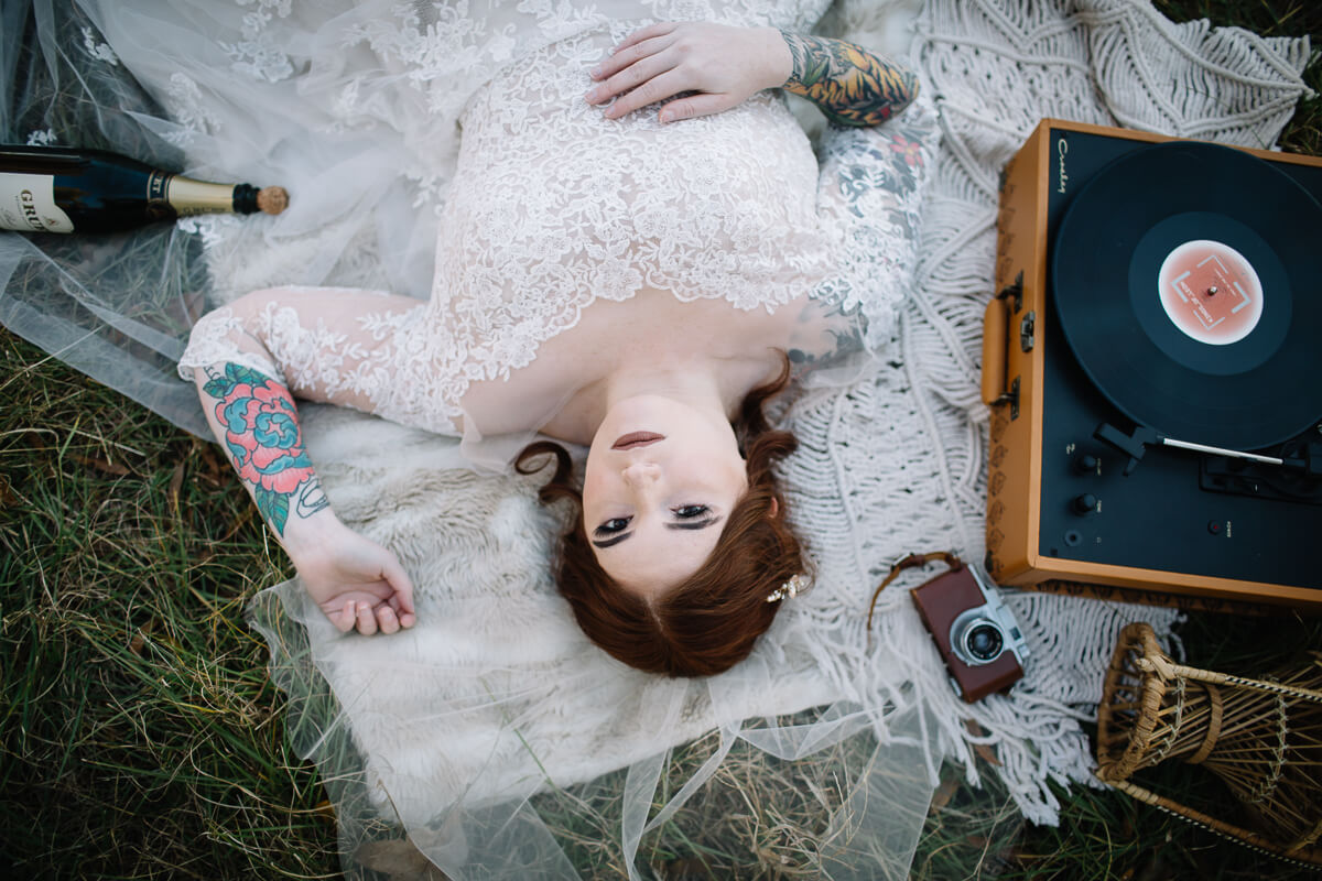 redhead bride in white laying on picnic blanket with record player antique camera and champagne bottle